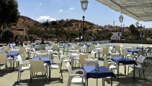 An outdoor seating area with tables and chairs, a covered section with hanging lanterns, and a view of a hilly landscape in the background.