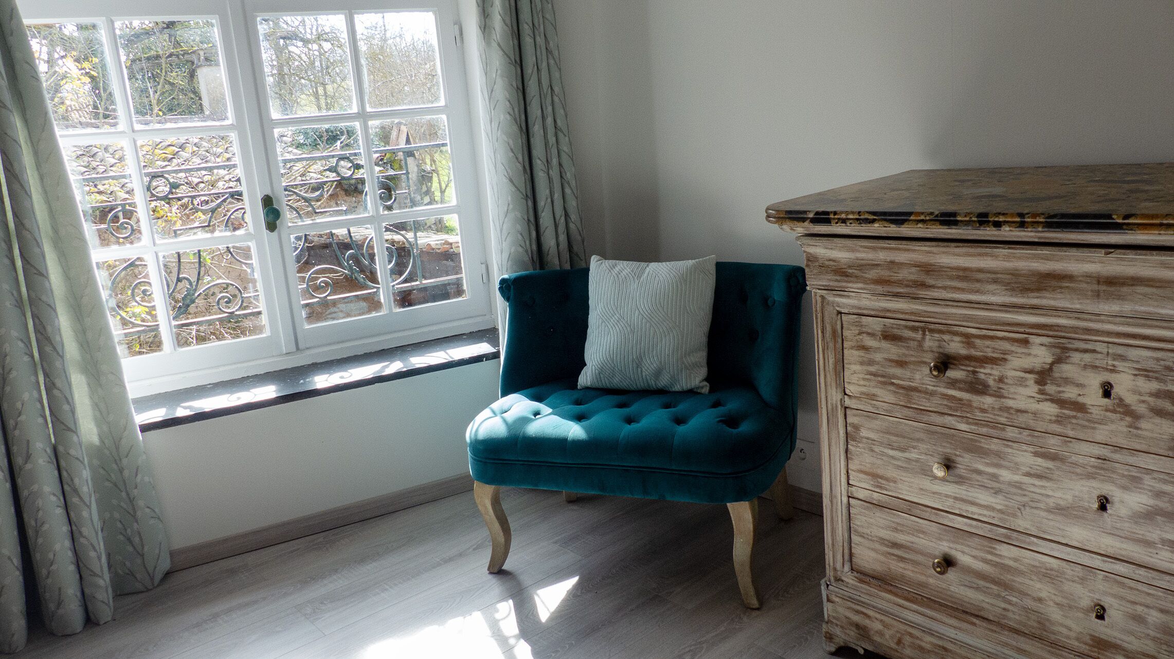 Une pièce comprenant un fauteuil recouvert d’un tissu de couleur turquoise, une commode en bois et une fenêtre donnant sur des arbres.