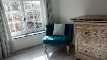 A room with a teal upholstered chair, a wooden dresser, and a window with a view of trees.