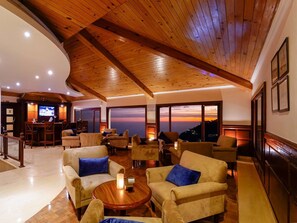 A spacious lobby with wooden ceiling beams, comfortable seating, a bar area, and large windows offering a view of the sunset. - Madhuban Sarovar Portico Mussoorie (Dehradun)