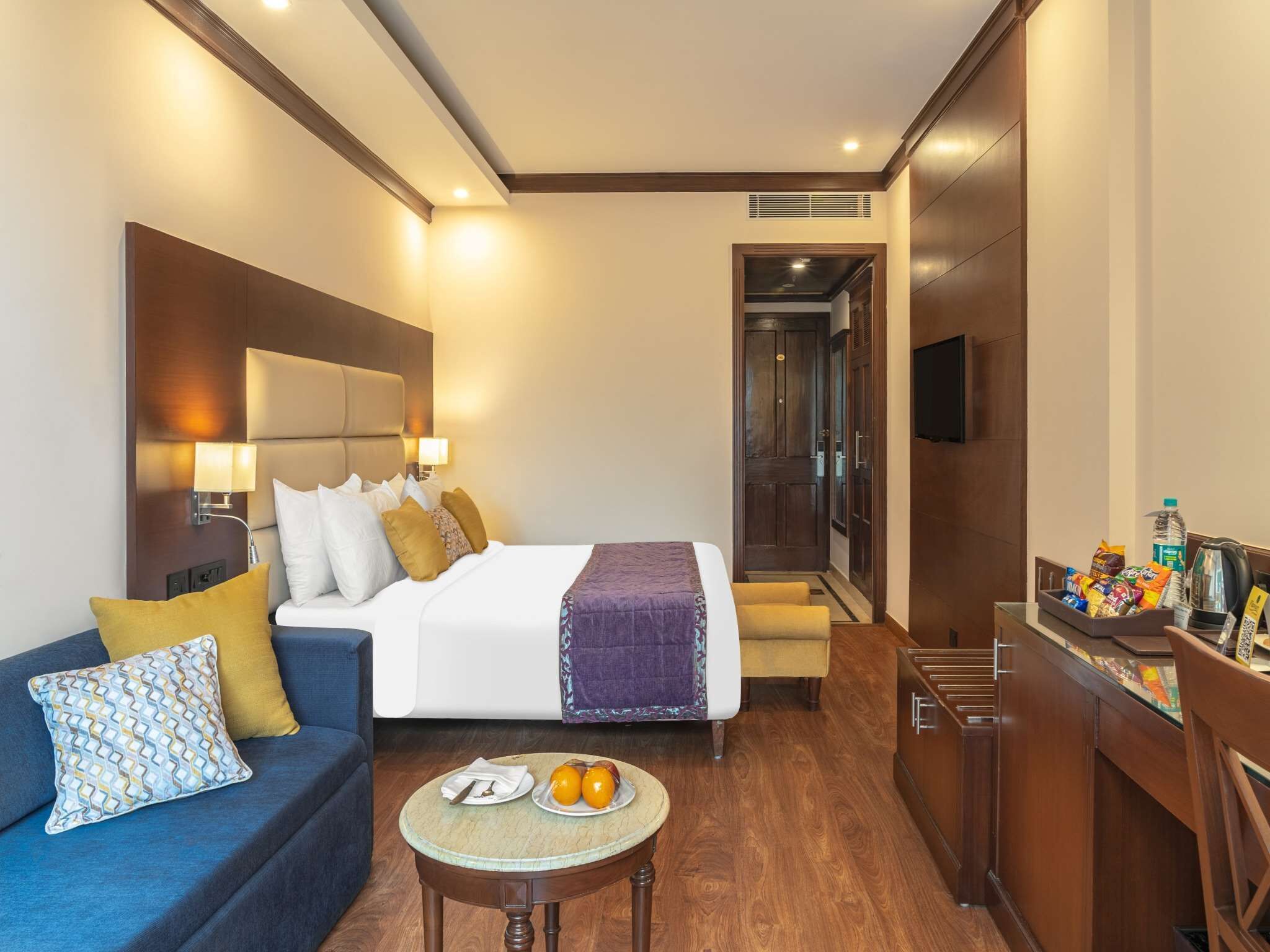 Una habitación de hotel con cama, sofá, una mesita con fruta, escritorio con bocadillos y televisor.