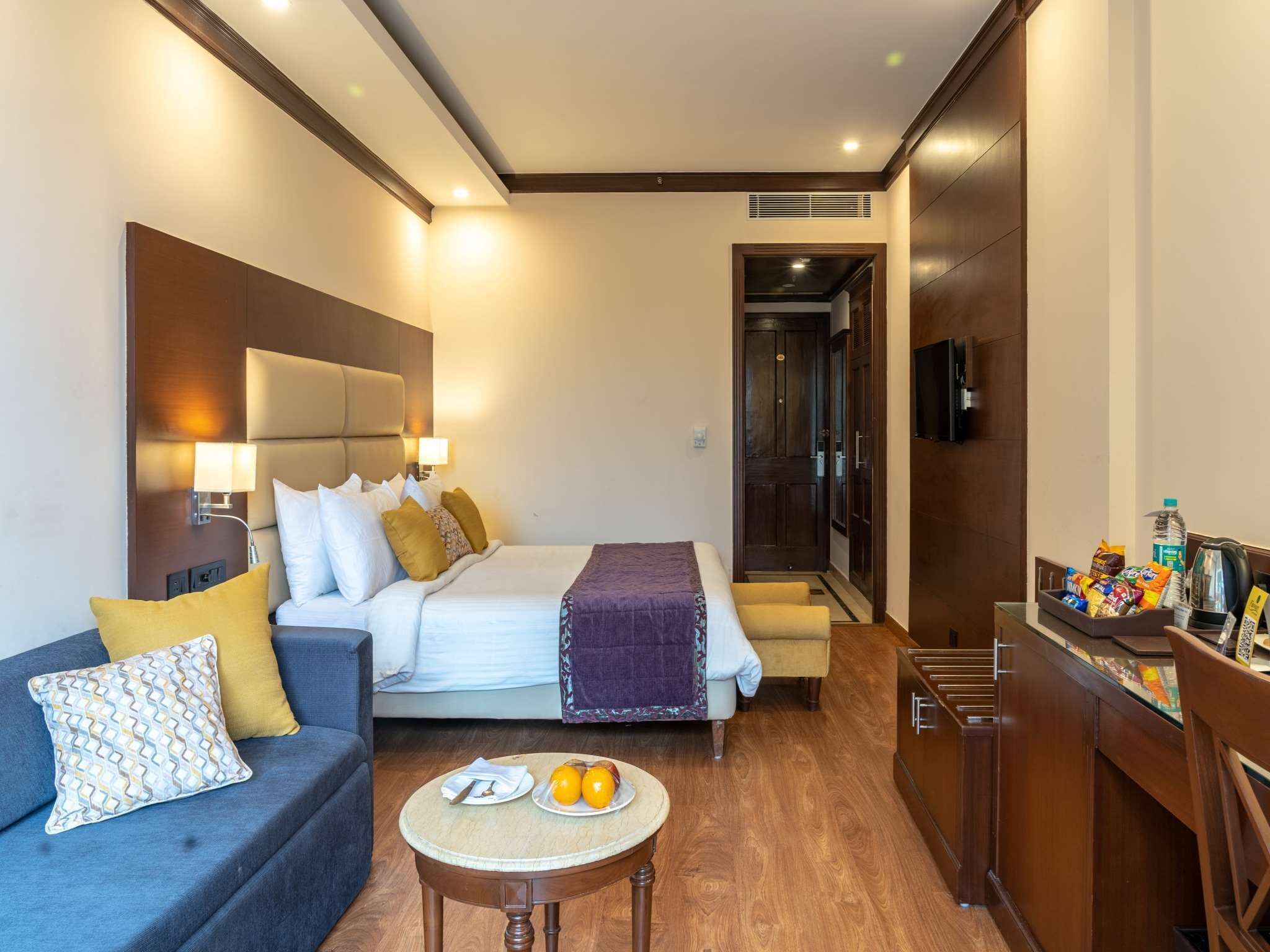 Una habitación de hotel con cama, sofá, una pequeña mesa con frutas y un minibar.