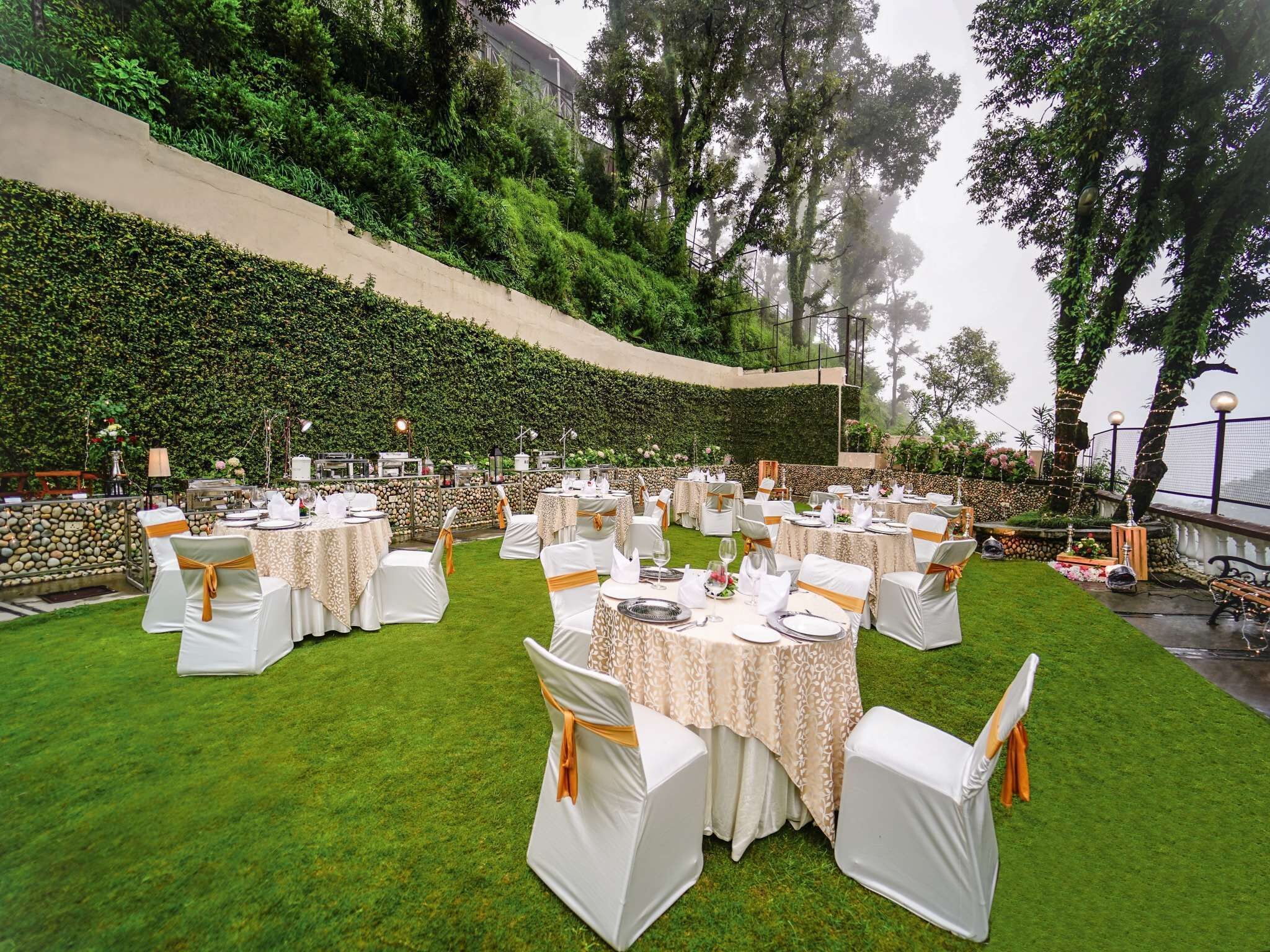 Un espacio para bodas al aire libre con mesas redondas y sillas blancas, frente a un fondo de pared de piedra y exuberante vegetación.