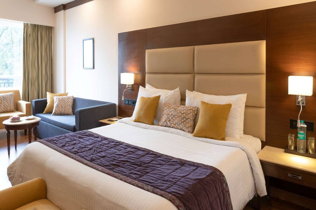 A hotel room with a large bed, a sofa, a chair, a bedside table with lamps, and a window with curtains.