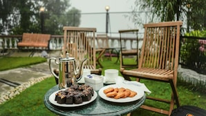 A table with a teapot, a plate of cakes, and a plate of cookies, set outdoors with chairs and a lamp in the background.
