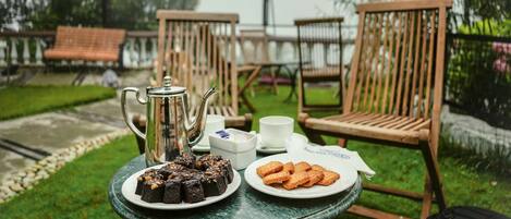 A table with a teapot, a plate of cakes, and a plate of cookies, set outdoors with chairs and a lamp in the background.