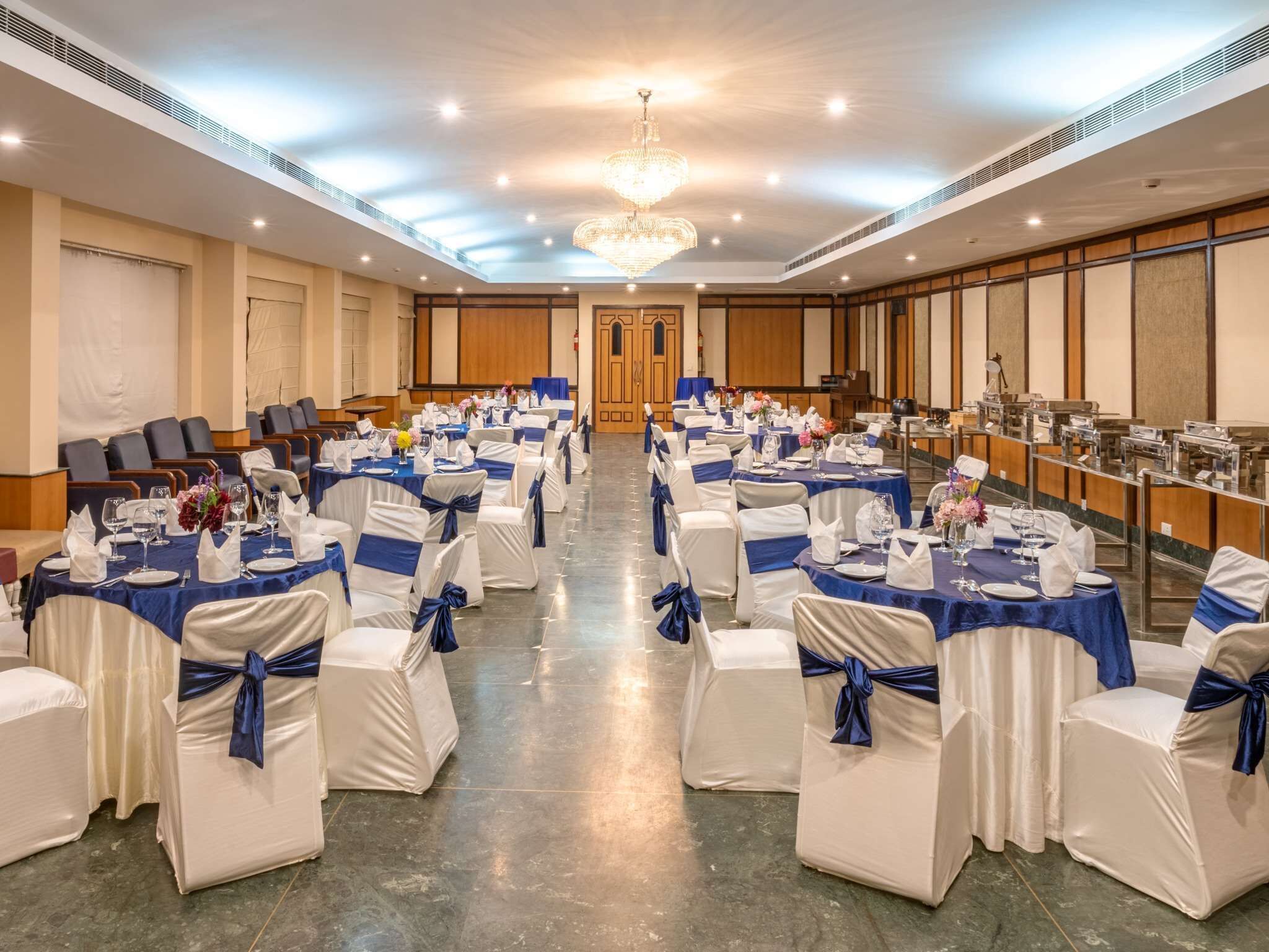 A banquet hall with rows of tables set for a formal event, featuring white chairs with blue sashes, a chandelier, and a polished floor.