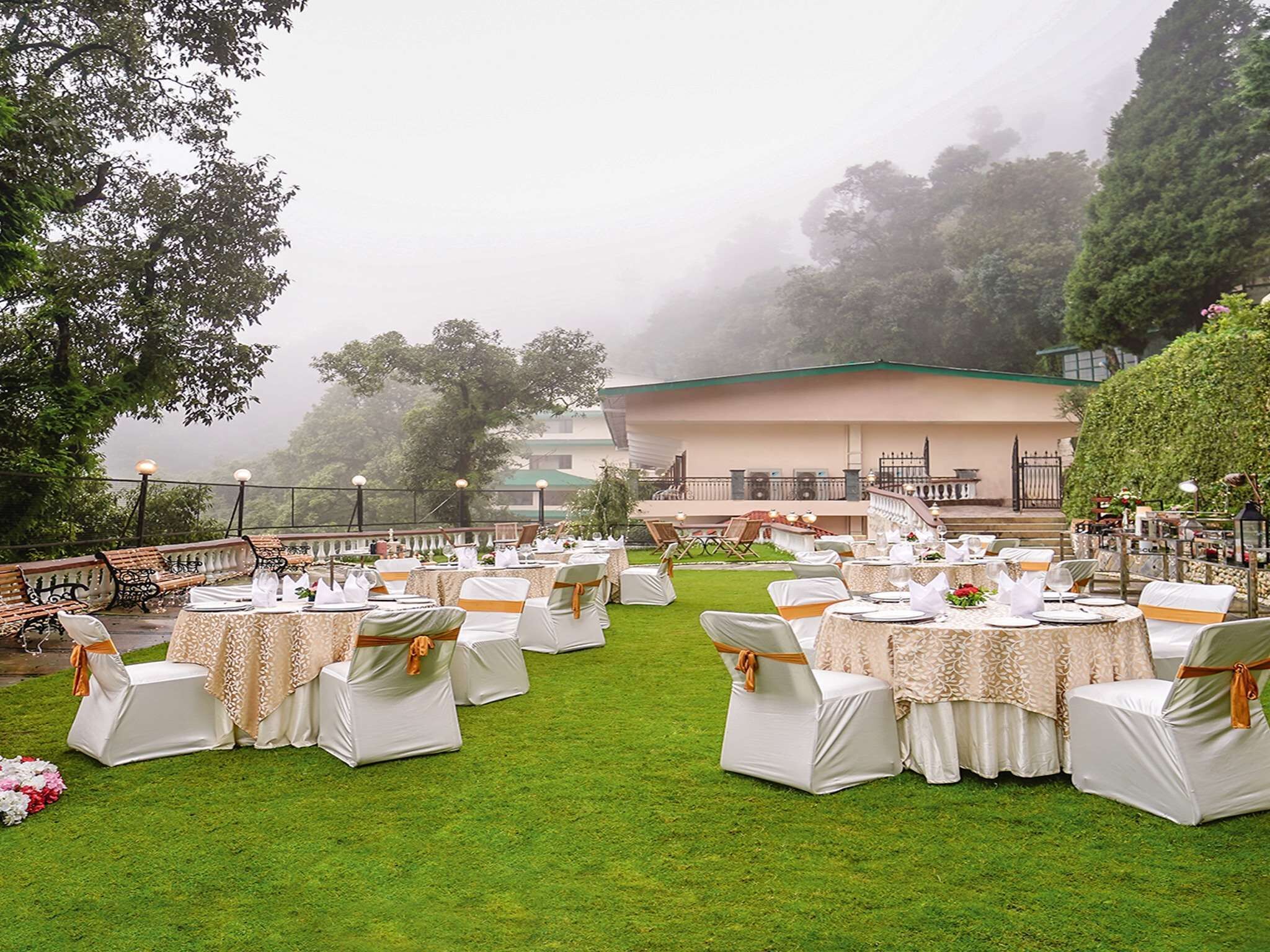 An outdoor wedding venue with tables and chairs set up on a grassy area, overlooking a building and trees.