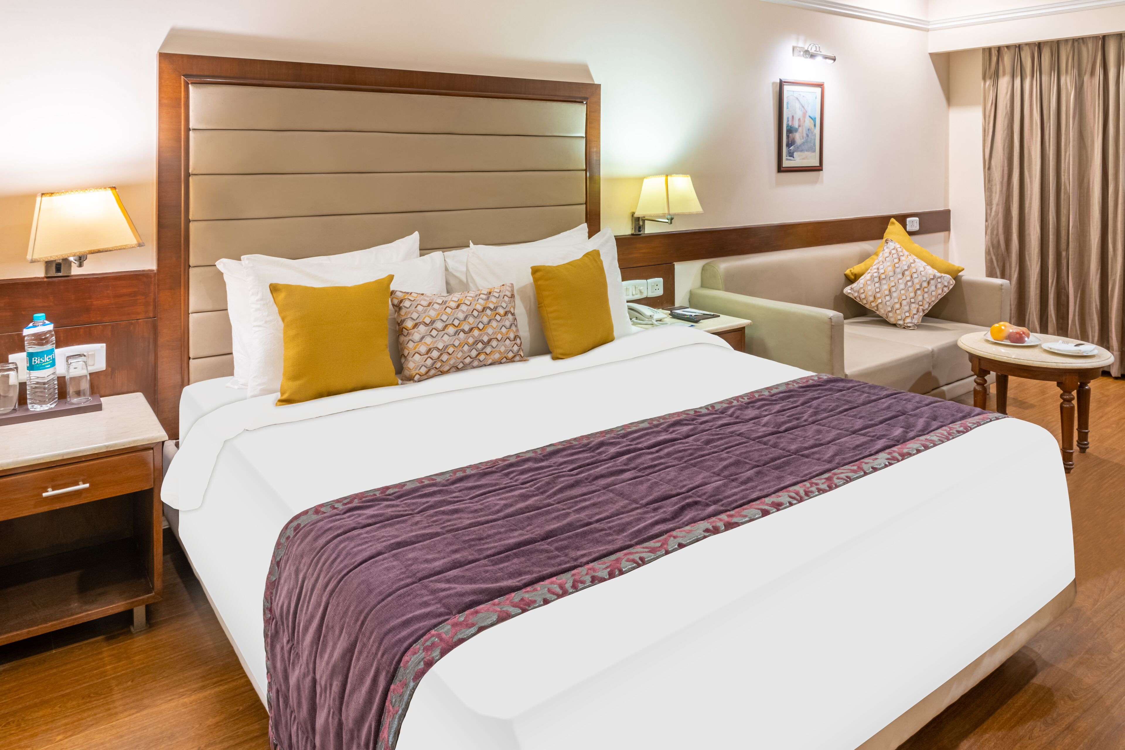 Een hotelkamer met een groot bed, een bank, een nachtkastje met lamp en een tafeltje met een schaal fruit.