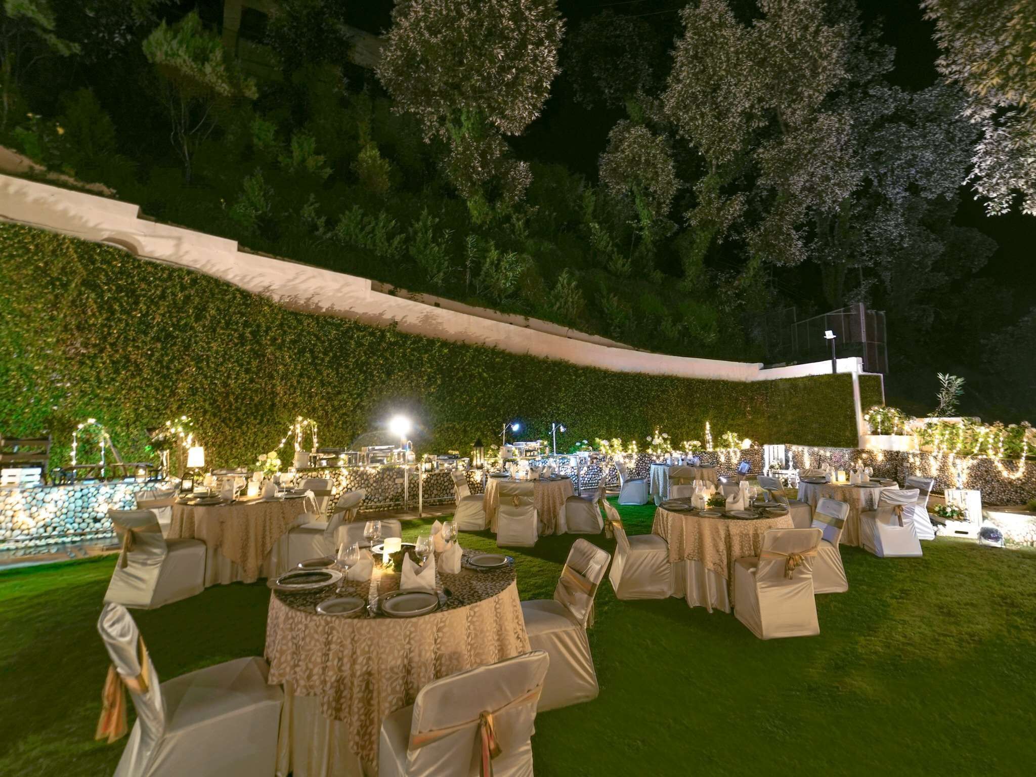 An outdoor event space with round tables and white chairs, surrounded by a lush green hedge and trees.
