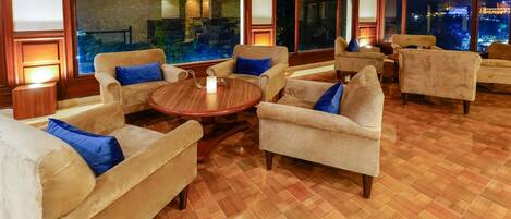 A spacious lounge area with multiple sofas and armchairs arranged around a central round table, large windows offering a view of the cityscape, and wooden flooring.