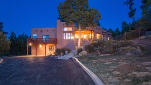 A modern two-story house with a balcony, a large tree in front, and a well-lit driveway.