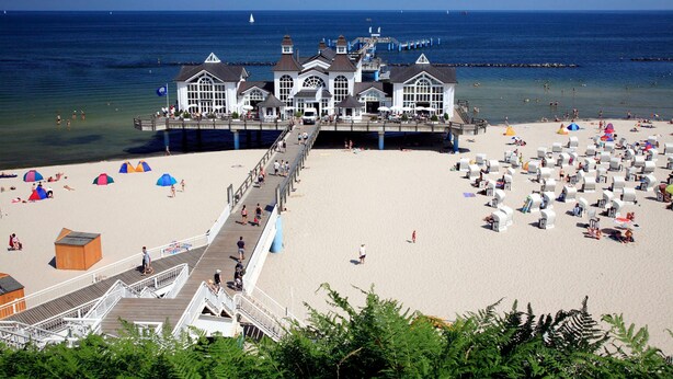 A pier leading to a white building with multiple windows and a balcony, overlooking a sandy beach with umbrellas and beach chairs. - Ferienwohnung mit Terrasse am Hafen in Seedorf 08 - 08 Ferienwohnung mit Terrasse am Hafen in (Sellin)