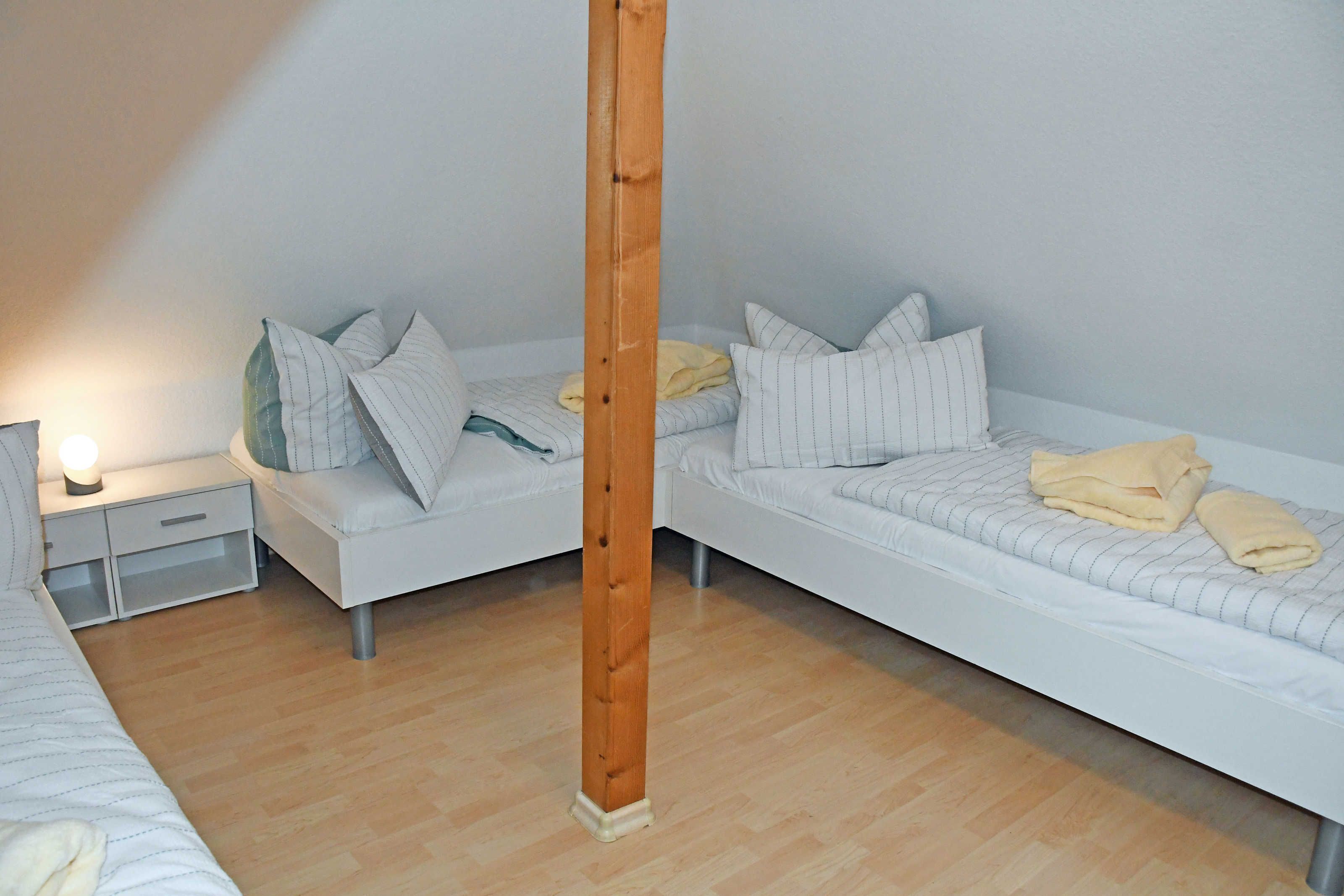 A room with two single beds, a wooden pillar, a nightstand with a lamp, and a small drawer unit.