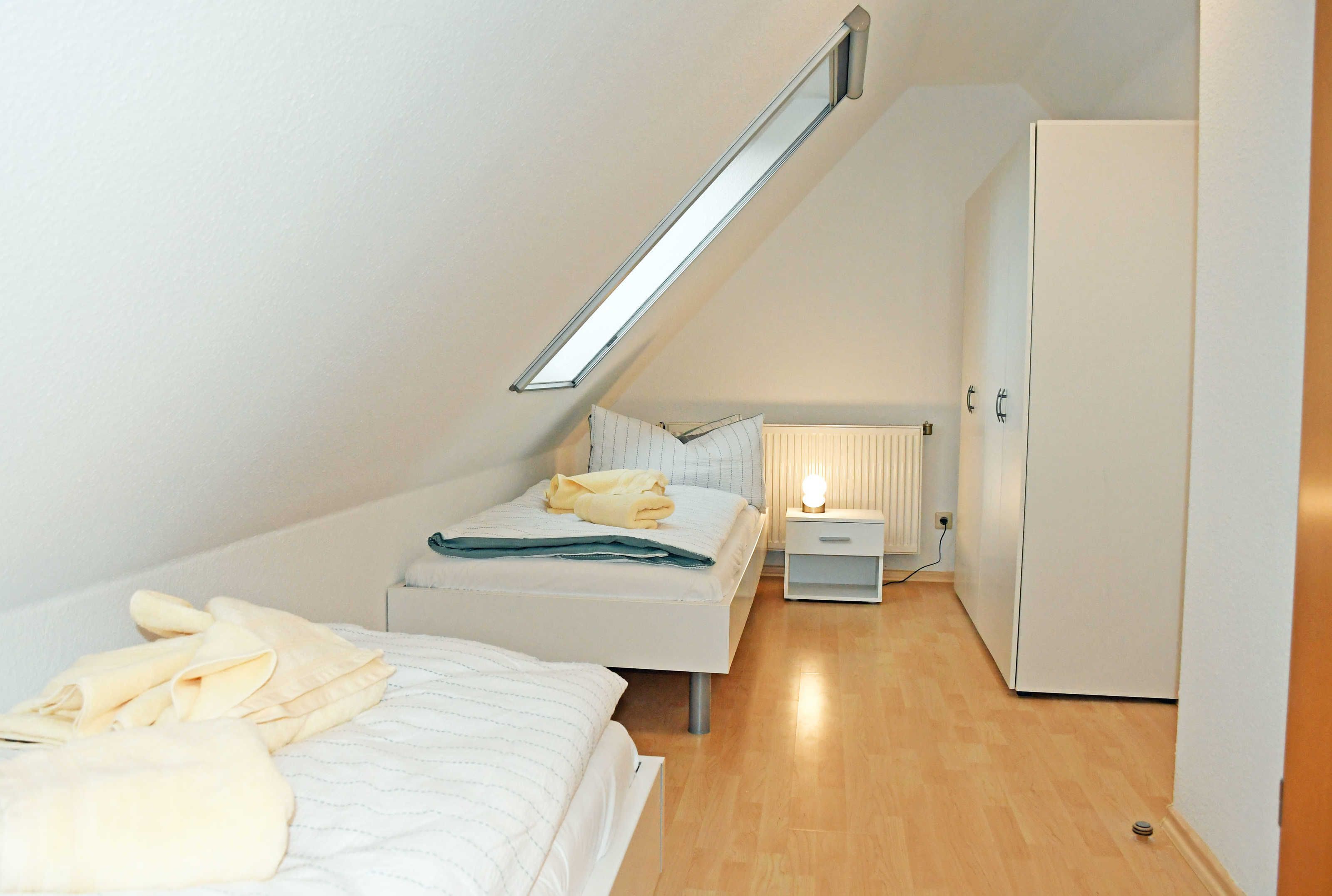 A small, well-lit bedroom with a sloped ceiling, a bed with white bedding, a bedside table with a lamp, and a wardrobe.