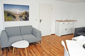 A living room with a grey sofa, a small white table, and a wooden floor. There is a white door with a cabinet above it.