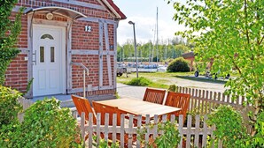 A brick building with a white door and the number 12 on it. There is a wooden table and chairs in front of the building, and a wooden fence partially obscures the view. - 03 Ferienwohnung m. Seeblick u. Terrasse am Hafen in Seedorf - Ferienwohnungen am Hafen in Seedorf (Sellin)