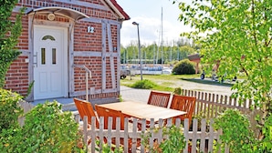 A brick building with a white door and the number 12 on it. There is a wooden table and chairs in front of the building, and a wooden fence partially obscures the view.