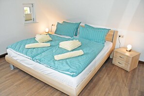 Cama bem arrumada com colcha listrada em azul e branco, dois travesseiros e duas toalhas enroladas. Tem uma mesa de cabeceira de madeira com abajur e uma tomada na parede.