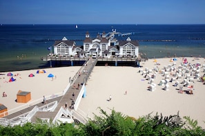 Eine Steg, der zu einem weißen Gebäude mit zahlreichen Fenstern führt, ein Sandstrand mit Sonnenschirmen und das Meer im Hintergrund.