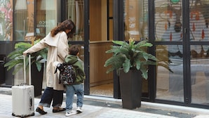 Una mujer y un niño con una maleta frente a un edificio con ventanas grandes y plantas en macetas.