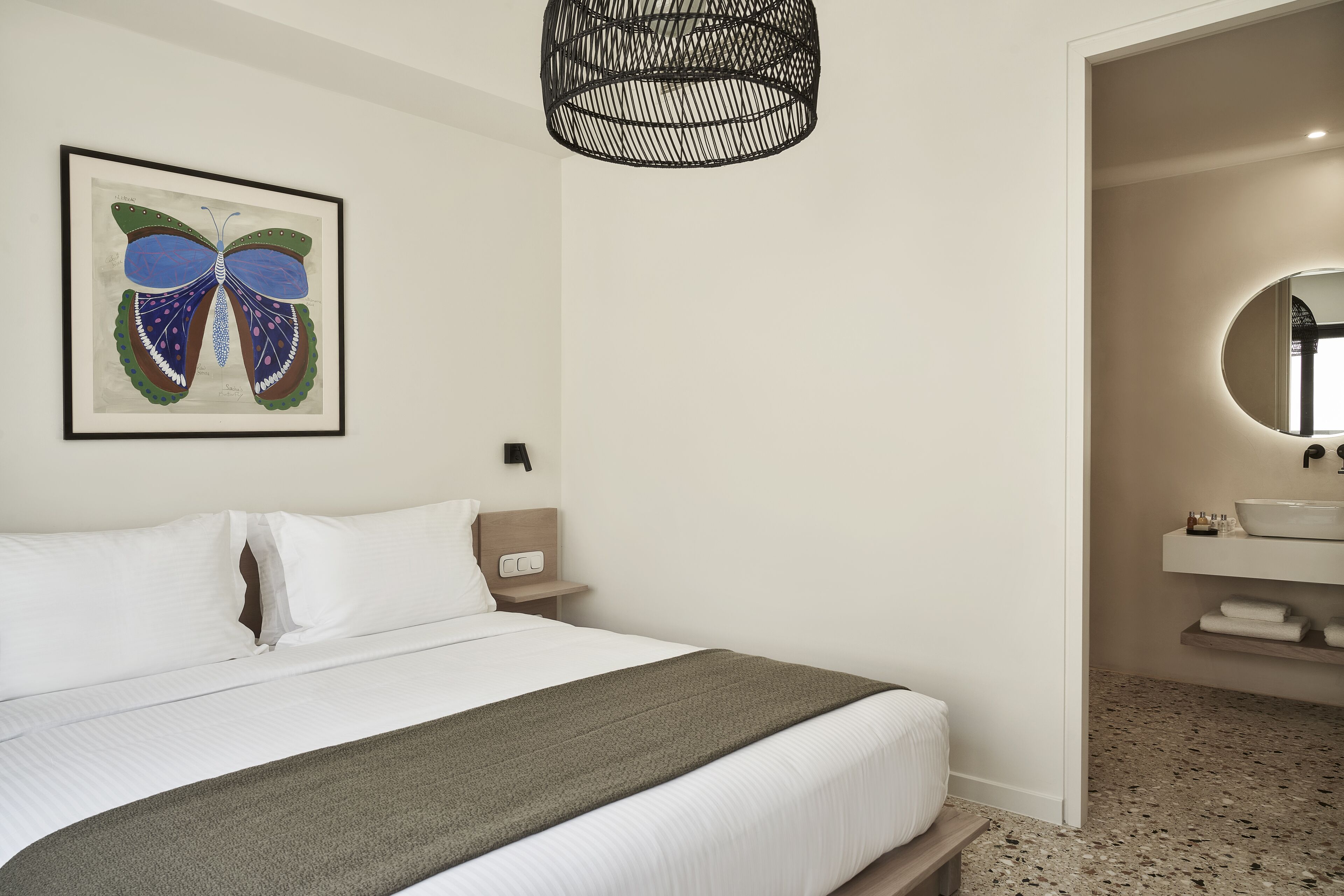 Quarto de hotel com cama, quadro de borboleta emoldurada, luminária pendente moderna, mesa de cabeceira e banheiro com espelho redondo e pia.