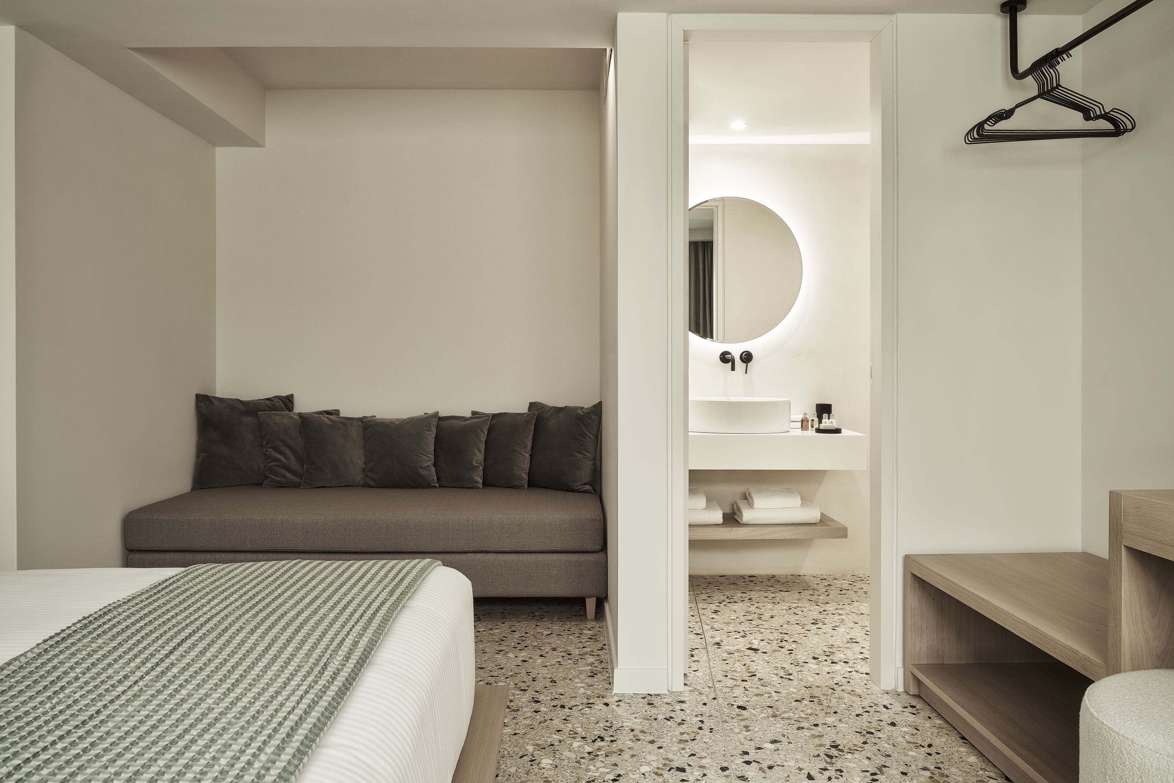 A modern hotel room with a bed, a sofa, a round mirror, a small sink, and a wooden shelf.
