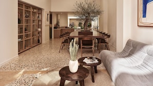 Amplia sala de estar con una mesa de comedor, un sofá y una mesa de centro. Se observa una estantería y una planta en maceta al frente.