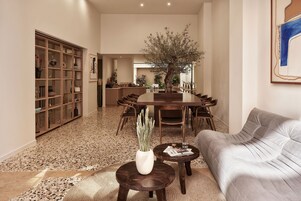 Uma sala ampla com uma mesa de jantar, um sofá e uma mesa de centro. Há uma estante de livros e um vaso de planta em primeiro plano.