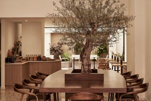 Uma sala de jantar com uma grande mesa de madeira rodeada por cadeiras, uma árvore central e um balcão com utensílios de cozinha.