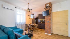 A modern living room with a blue sofa, a dining area with a table and chairs, a kitchen with a TV mounted on a brick wall, and a wooden door. - 4*Flat in the Heart of the City, Onsite parking, Wifi (Split)