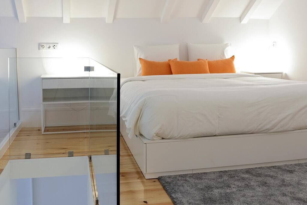 Une chambre moderne avec une table de chevet en verre, un lit blanc agrémenté d’oreillers orange et un tapis gris.