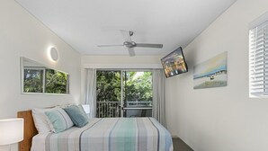Ein Schlafzimmer mit Bett, Nachttisch, Sessel, Deckenventilator, Fernseher und einem Fenster mit Vorhängen.