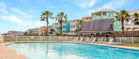 Una piscina con sillones de descanso y una zona sombreada, rodeada de edificios coloridos y palmeras.