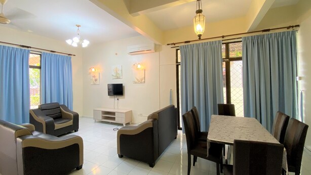 A spacious living area with a sofa set, a dining table, and chairs. There is a flat-screen TV mounted on the wall, and the room has a ceiling fan.
