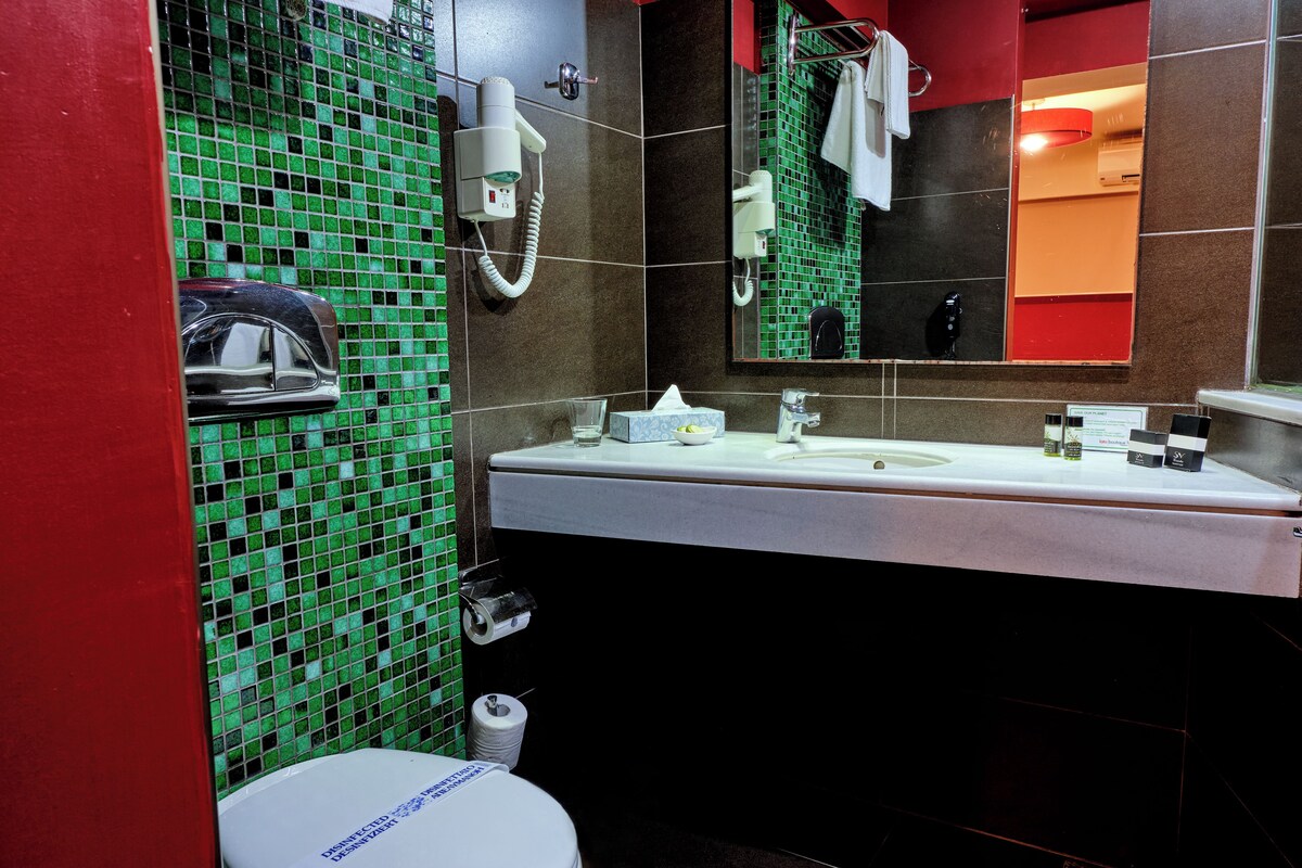 Un baño con un lavamanos blanco, un espejo, un muro cubierto de azulejos verdes, un inodoro blanco y una pared roja.