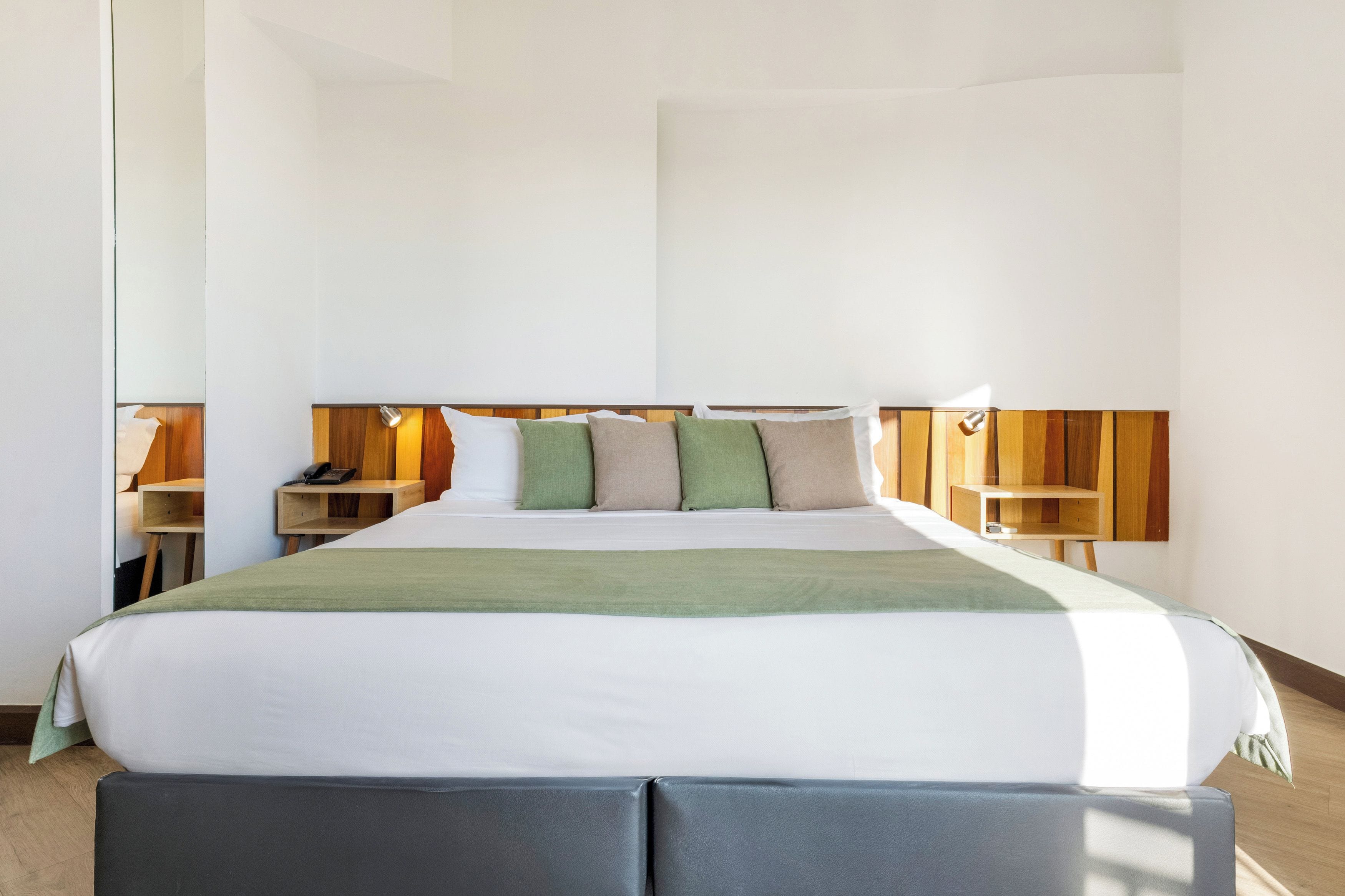 Une chambre d’hôtel moderne avec un grand lit, une tête de lit en bois, des tables de chevet avec des lampes et un plaid vert.