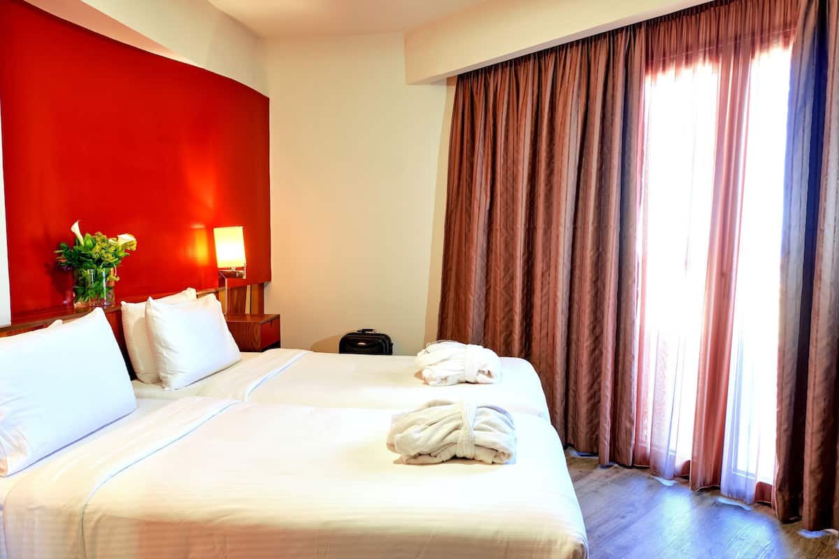 A hotel room with two beds, a red accent wall, a nightstand with a lamp, a suitcase, and a window with curtains.