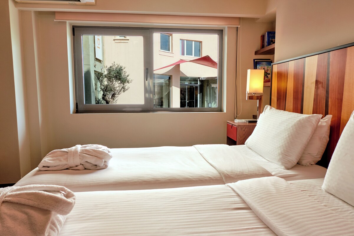 A hotel room with two beds, a window with a view of buildings, a bedside table with a lamp, and a wall-mounted shelf with books.