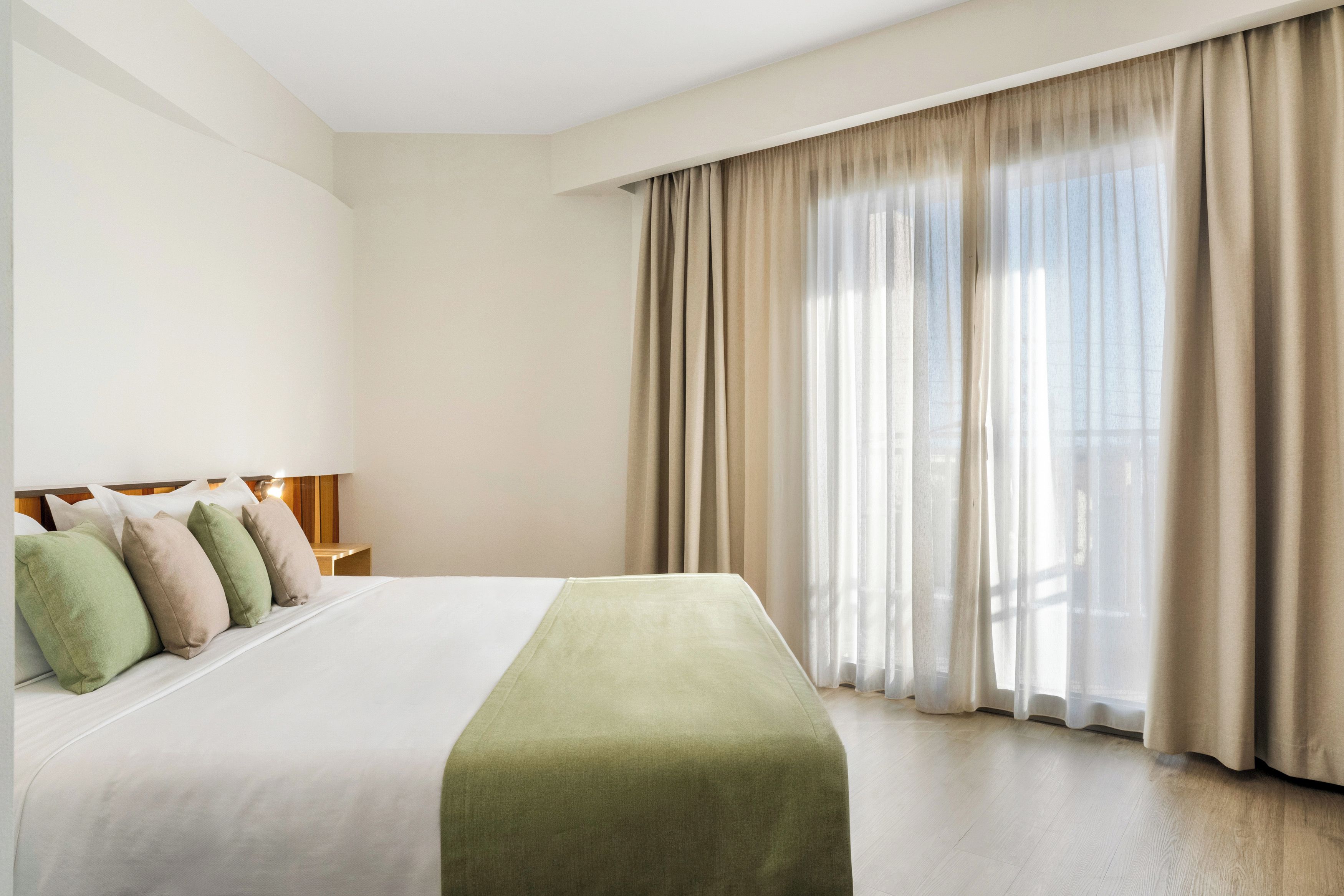 A hotel room with a large bed, a nightstand with a lamp, and a view through sheer curtains.