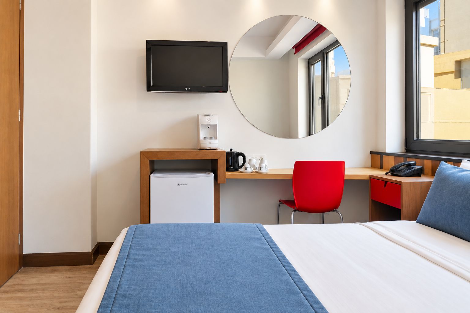 A hotel room with a bed, a desk with a coffee maker, a red chair, a small refrigerator, a telephone, and a television mounted on the wall.
