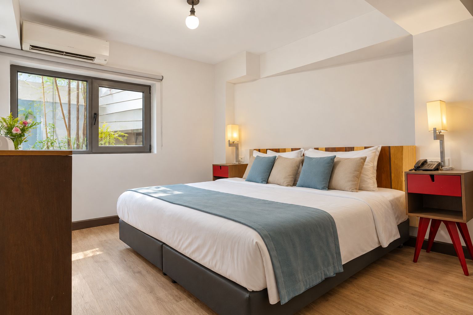 A modern hotel room with a large bed, bedside tables, and a window with a view of greenery.