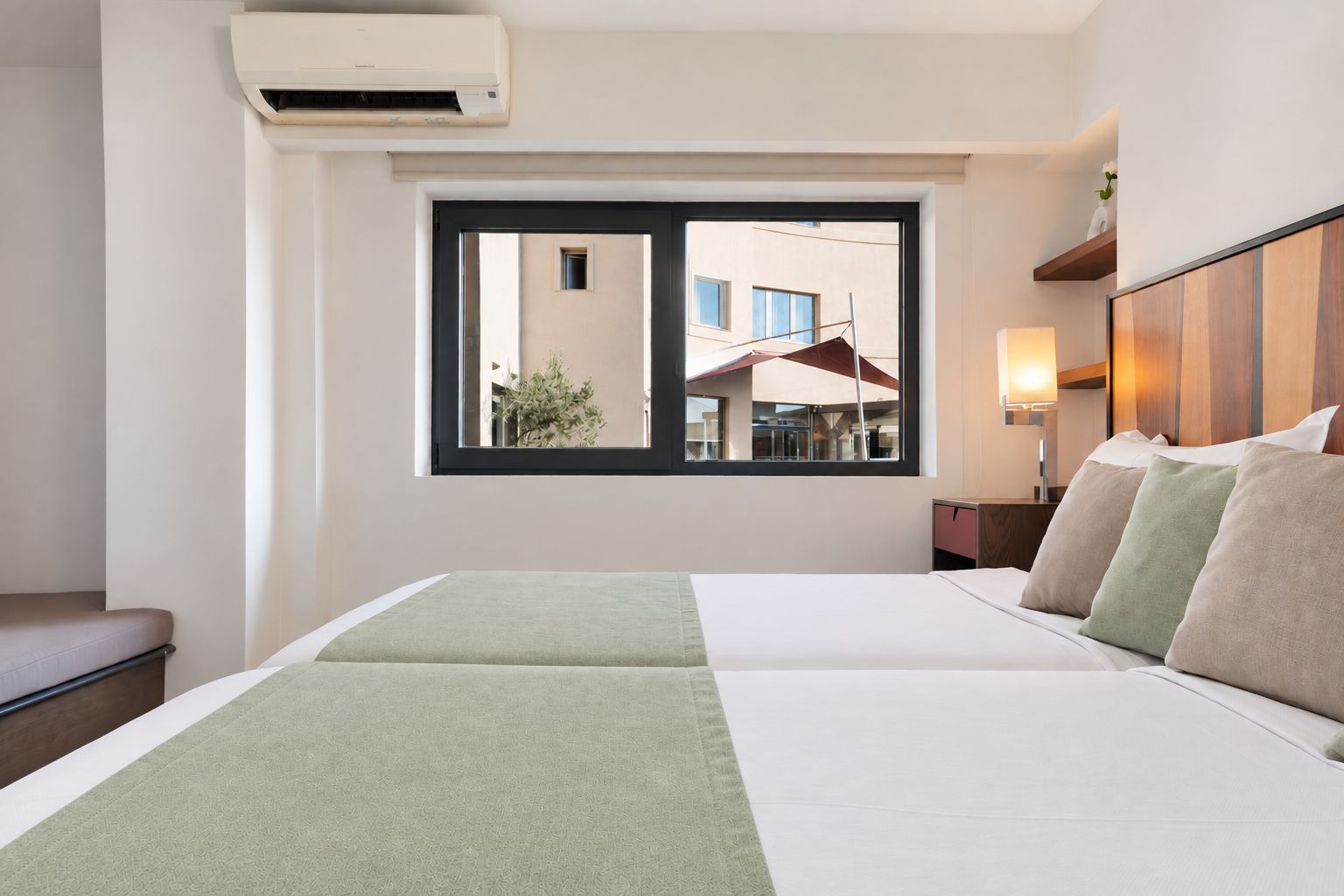 Habitación de hotel con cama, ventana, aire acondicionado de pared y mesita de noche con lámpara.