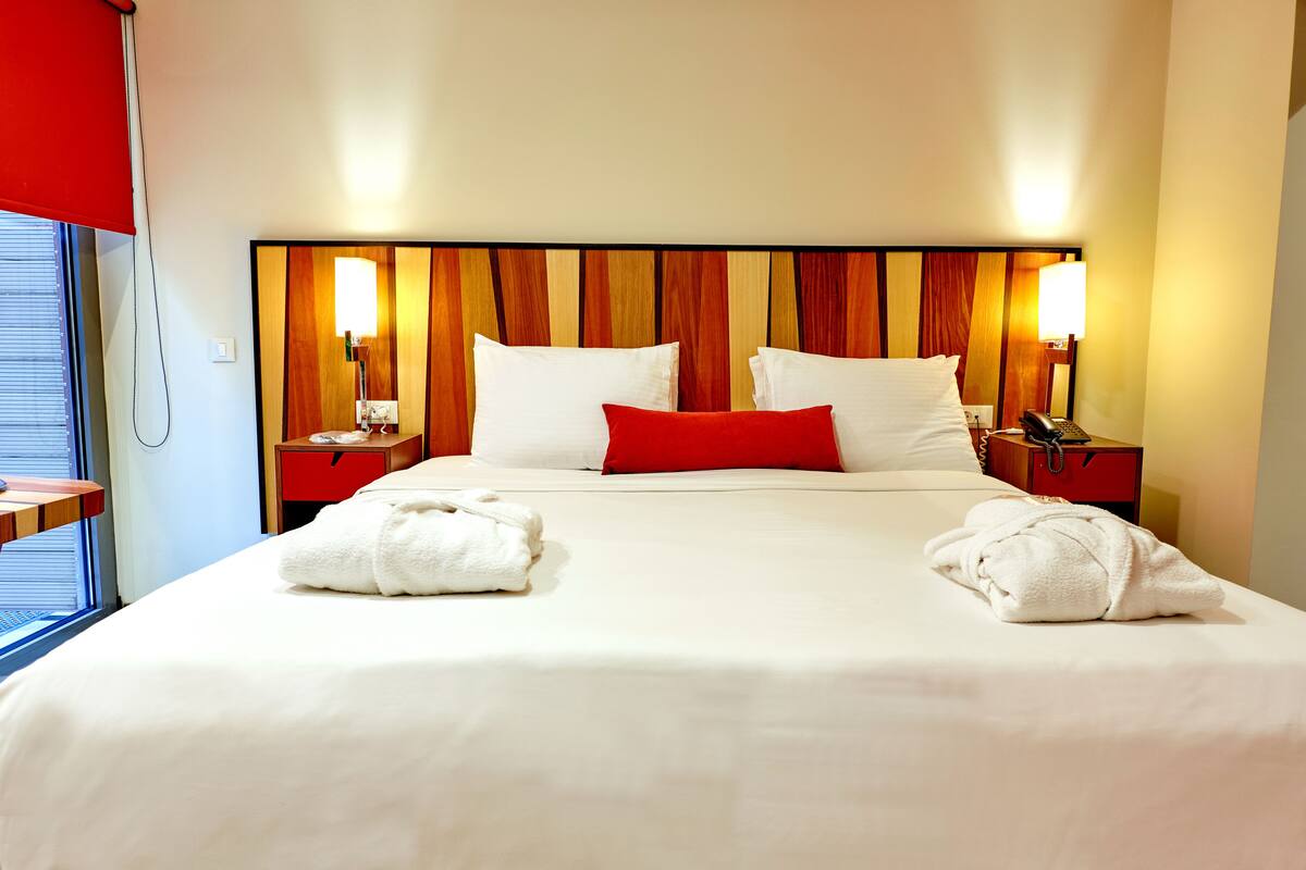 A hotel room with a large bed, two bedside tables, a red pillow, white pillows, two lamps, and two towels on the bed.