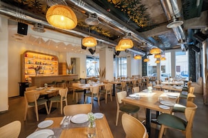 Un restaurante con mesas y sillas de madera, lámparas colgantes y una zona de bar con estantes llenos de botellas.