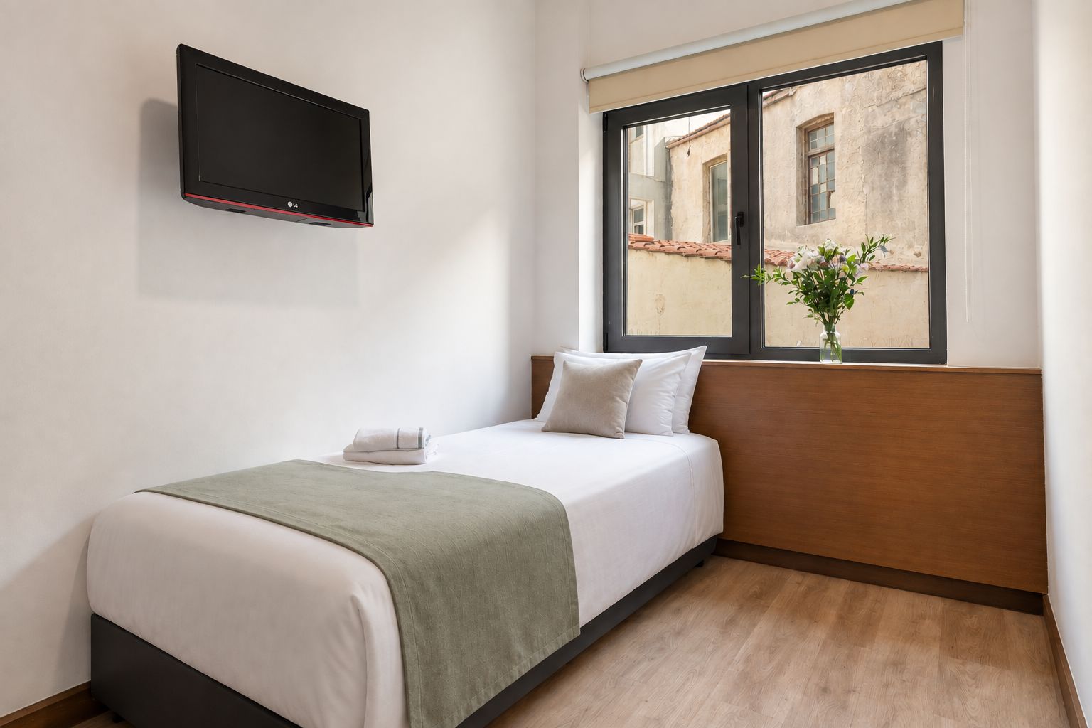 A single bed with a green blanket, white pillows, and a flat-screen TV mounted on the wall.