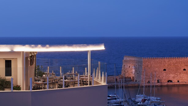 A rooftop terrace with tables and chairs, overlooking a body of water and a historic stone wall. - Lato Annex Boutique Rooms (Heraklion)