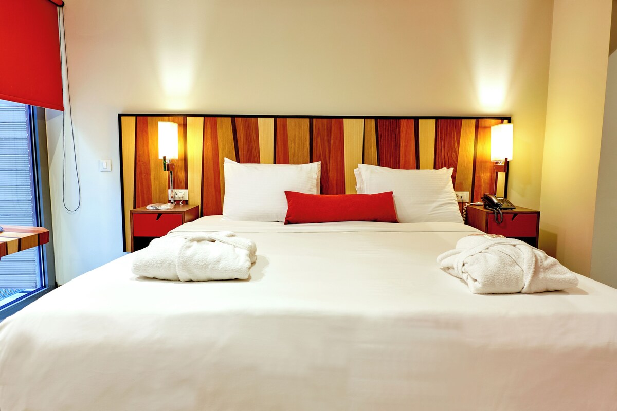 Habitación de hotel con una cama grande, dos mesitas de noche, un almohadón rojo, toallas blancas y un cabecero de madera.