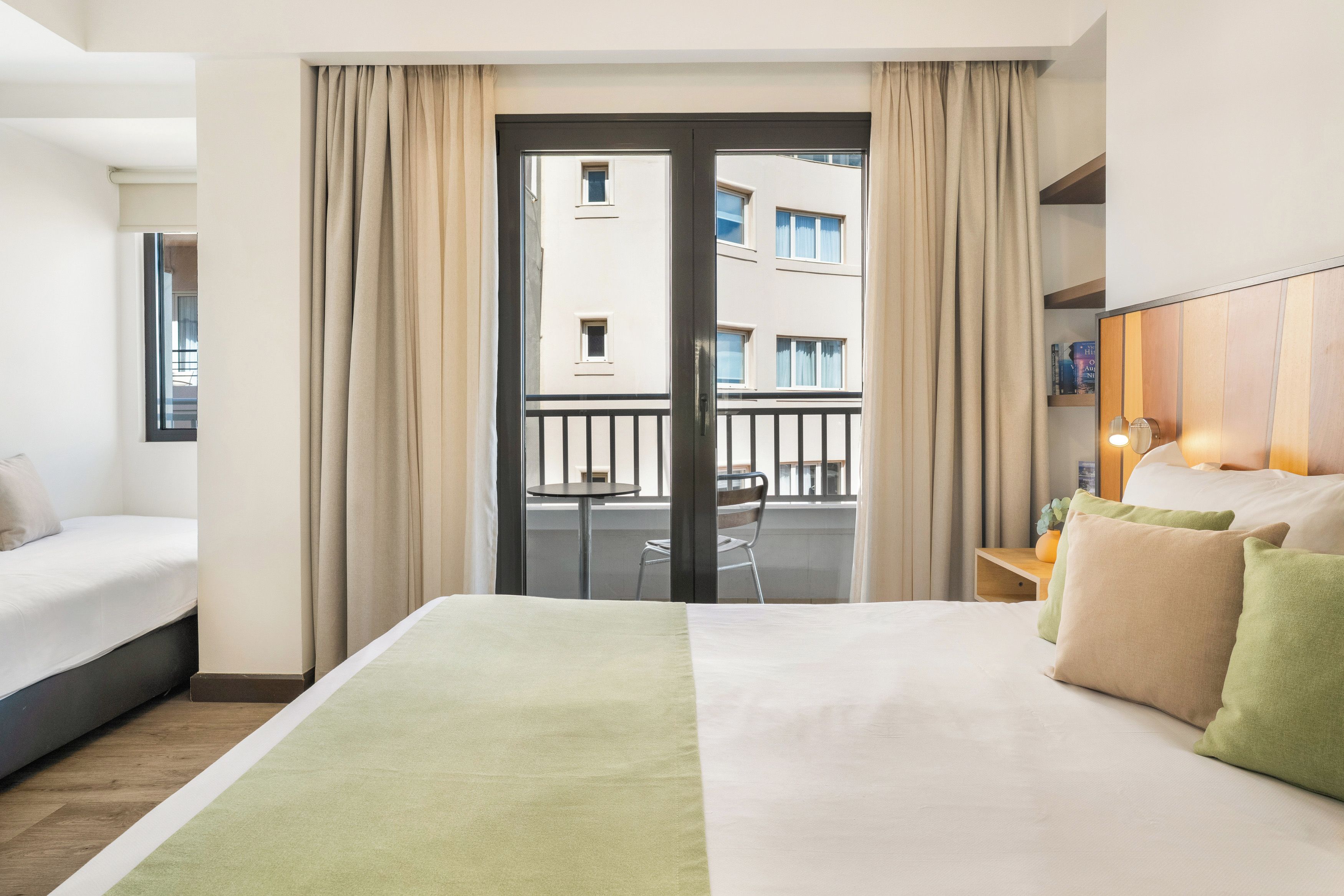 A hotel room with a bed, a small table, a chair, and a view of the outside building through a glass door.