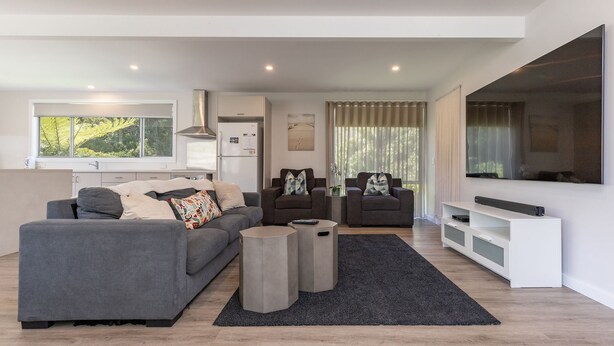 Ein modernes Wohnzimmer mit grauem Sofa, zwei Sesseln, einem Couchtisch und einem Fernseher. Es gibt eine Küchenzeile mit Kühlschrank und einem Fenster mit Blick auf Grünflächen.